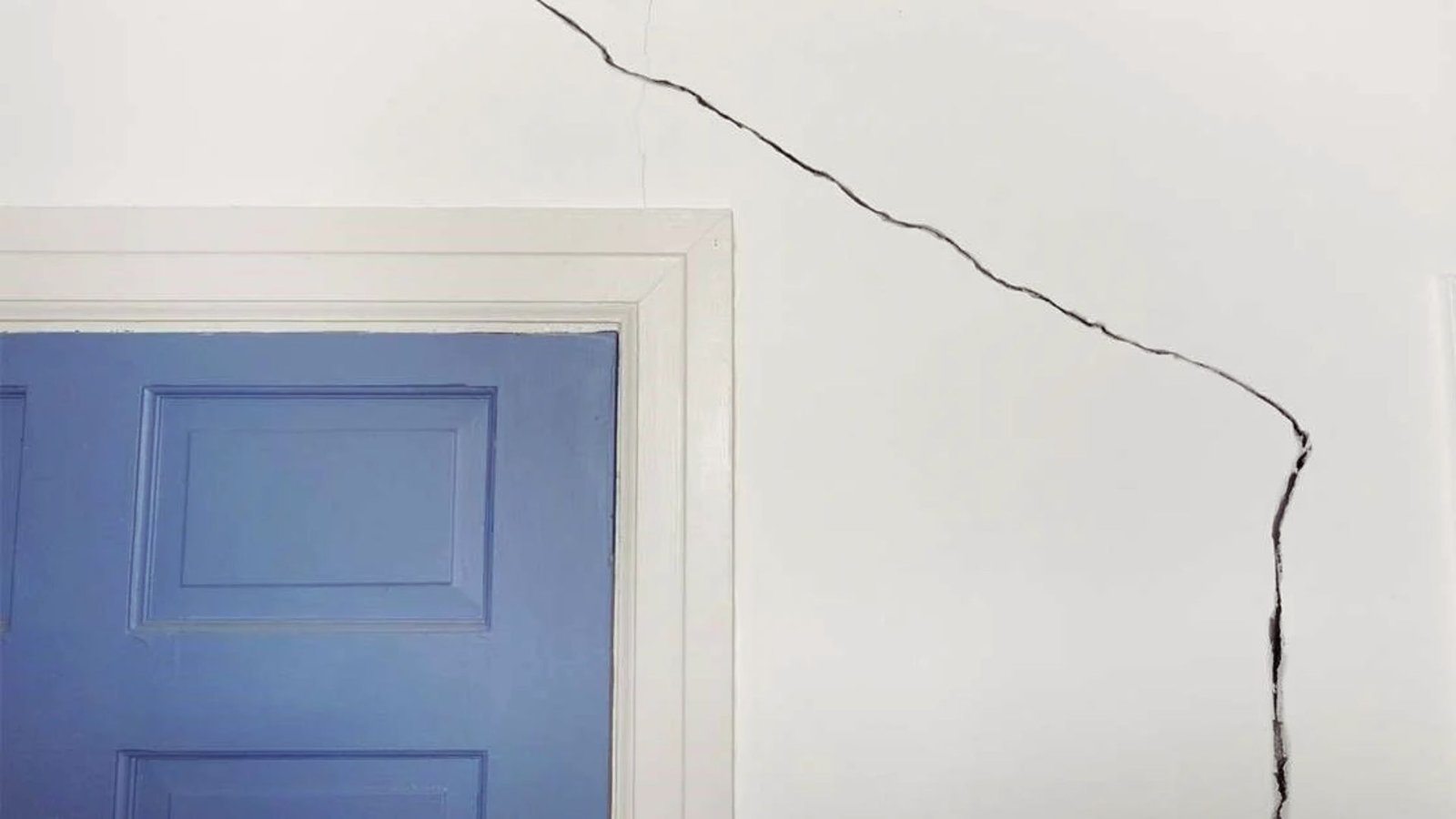 A crack in the wall of a room beside a blue door, highlighting wear and tear in the interior space.