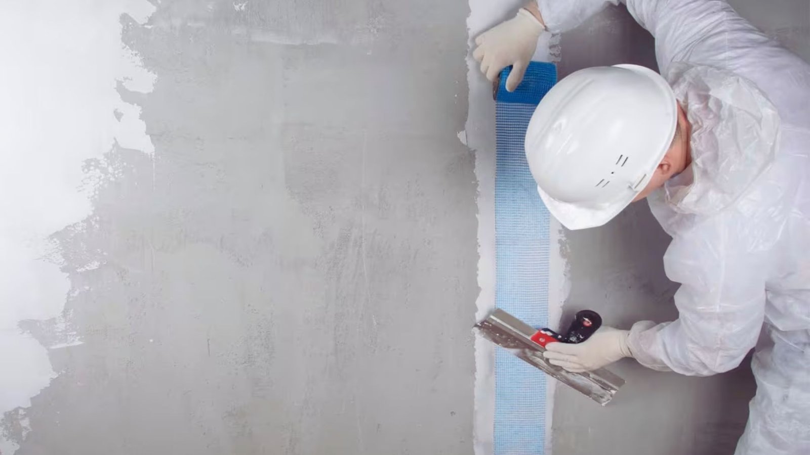 A man in a white suit and hard hat paints a wall, focused on his task with a paintbrush in hand.